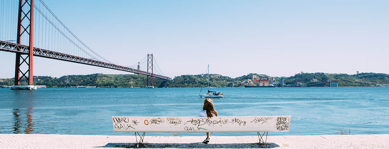 femme de dos assise sur un grand banc blanc face au fleuve Tage, vu sur la rive en face de la ville de Lisbonne et sur le pont rouge du 25 avril